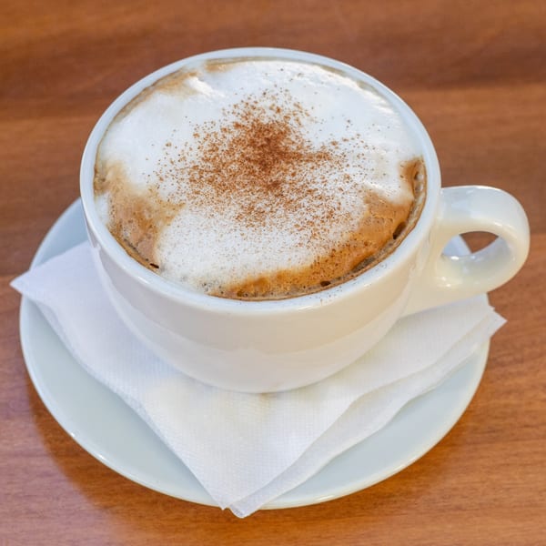 A white mug of cappuccino with frothy milk and a sprinkle of cinnamon on a wooden table.