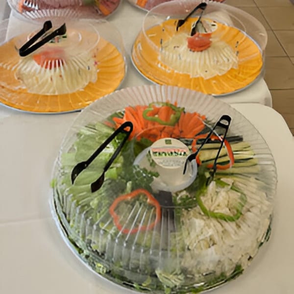 Three deli platters with salads and vegetables, covered with clear plastic lids and serving tongs.
