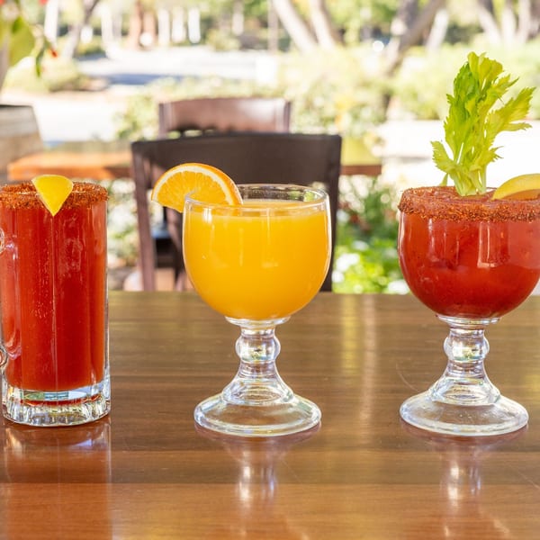 Three drinks on a wooden table: two Bloody Marys with garnishes and one orange juice.
