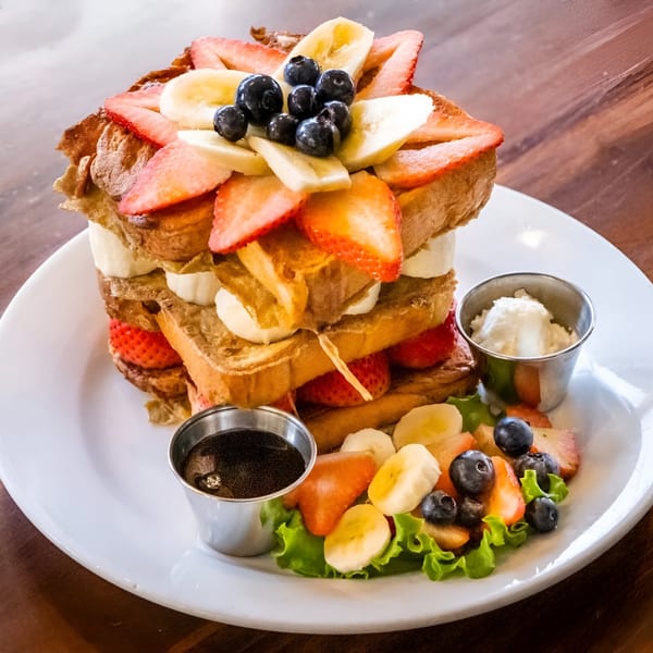 Stacked French toast with strawberries, blueberries, and bananas, served with syrup and whipped cream.