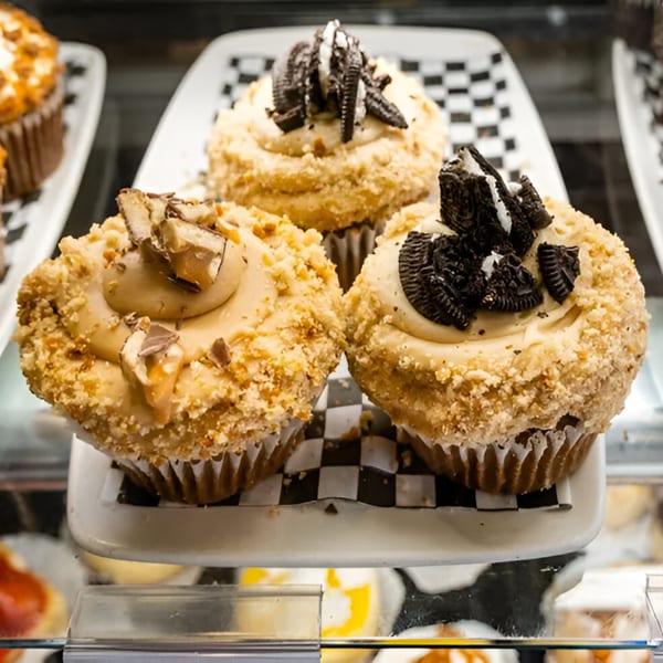 Three gourmet cupcakes, two with Oreo crumbles and one with peanut butter candy, on a checkered plate.