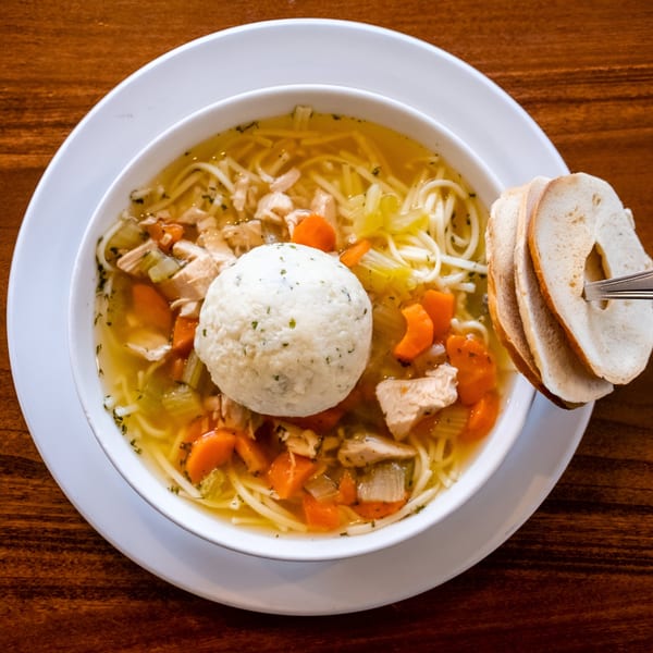 A bowl of Pickles Deli's matzo ball soup with noodles, chicken, carrots, and celery, served with bread.