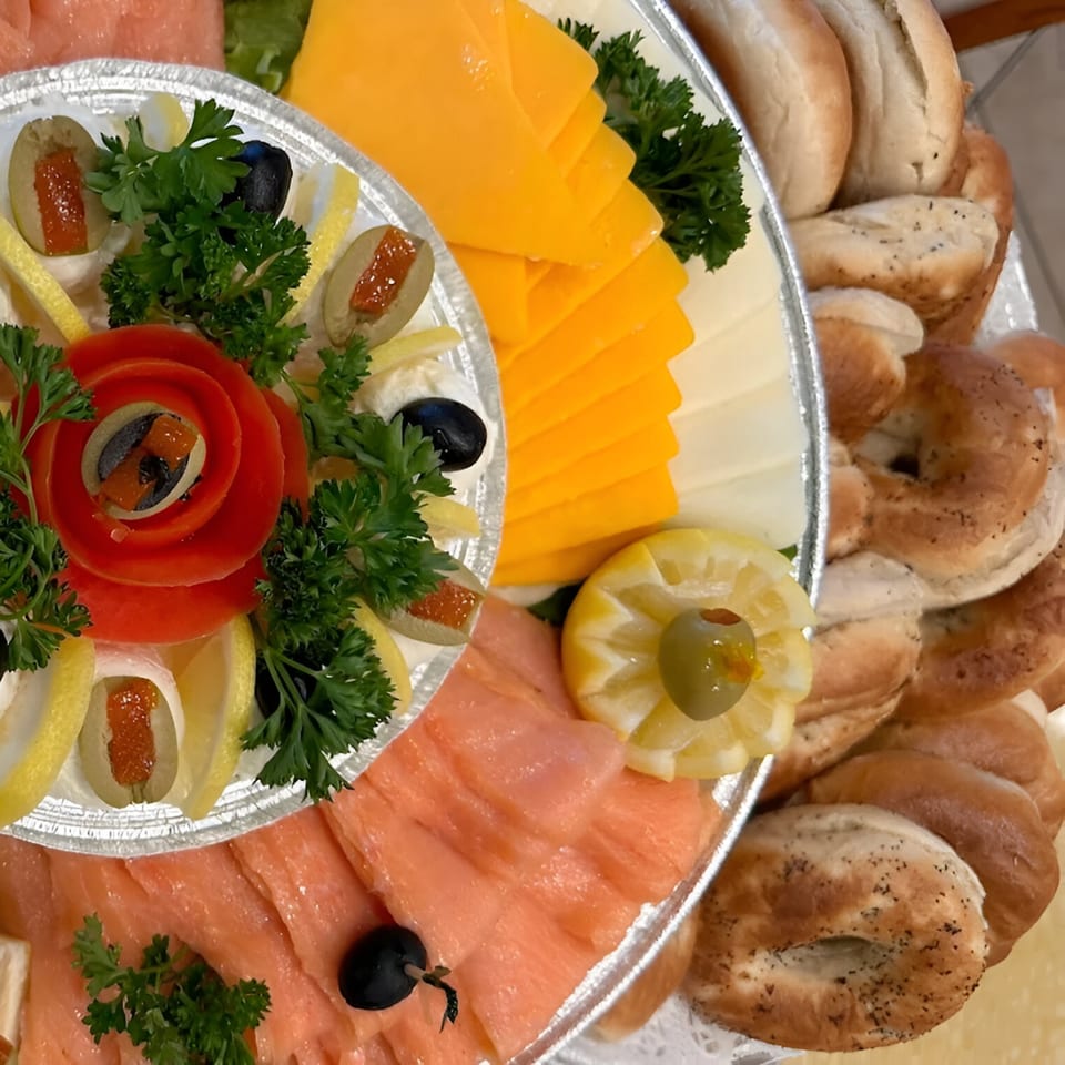 A deli platter with smoked salmon, sliced cheese, bagels, olives, lemons, and parsley.