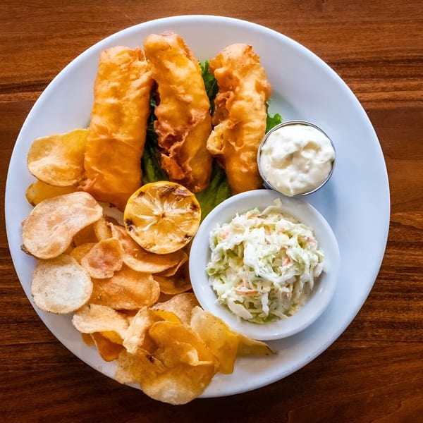 A white plate holds golden-brown fish and chips, coleslaw, tartar sauce, and a grilled lemon.