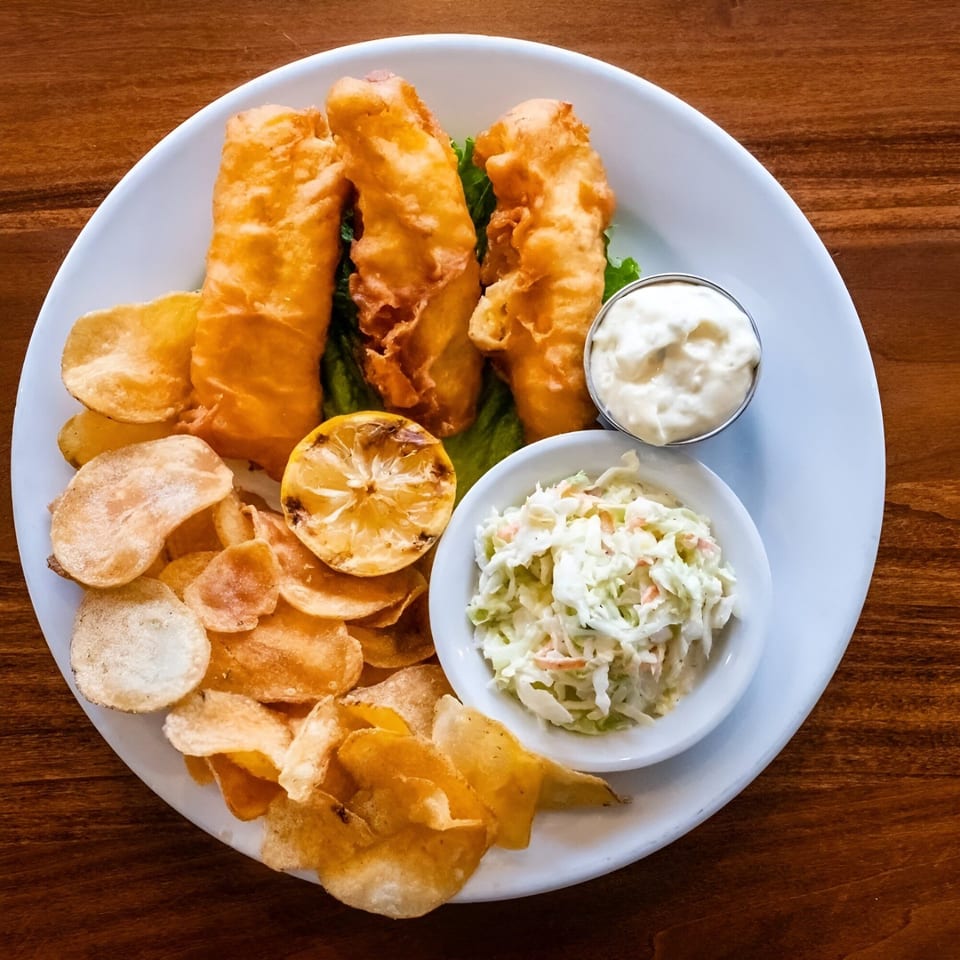 A white plate holds golden-brown fish and chips, coleslaw, tartar sauce, and a grilled lemon.